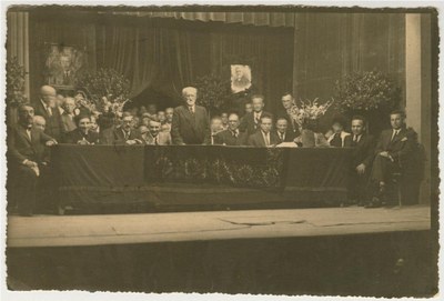 Simon Dubnow (standing, center) at the 1st international YIVO conference, Vilna, Poland. Other historians and notables on the stage and dais include (seated, from left) Joseph Tshernikhov, Elias Tcherikower, Yankev Botoshansky; (behind Botoshansky’s left shoulder) Zelig Kalmanovitch; (to Dubnow’s right) Ignacy Schiper; (behind Dubnow’s right shoulder) Rafail Abramovich; (seated, first to fourth from right) Yudl Mark, Yankev Shatzky, Max Weinreich, Zalmen Reyzen; (standing behind Weinreich), Nakhman Meisel. The portrait of Tsemaḥ Szabad (right) is draped in black in commemoration of his death, which had occurred a few months earlier.
